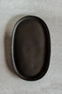 An oval black ceramic serving platter with a matte finish, photographed from directly above on a textured light gray surface.